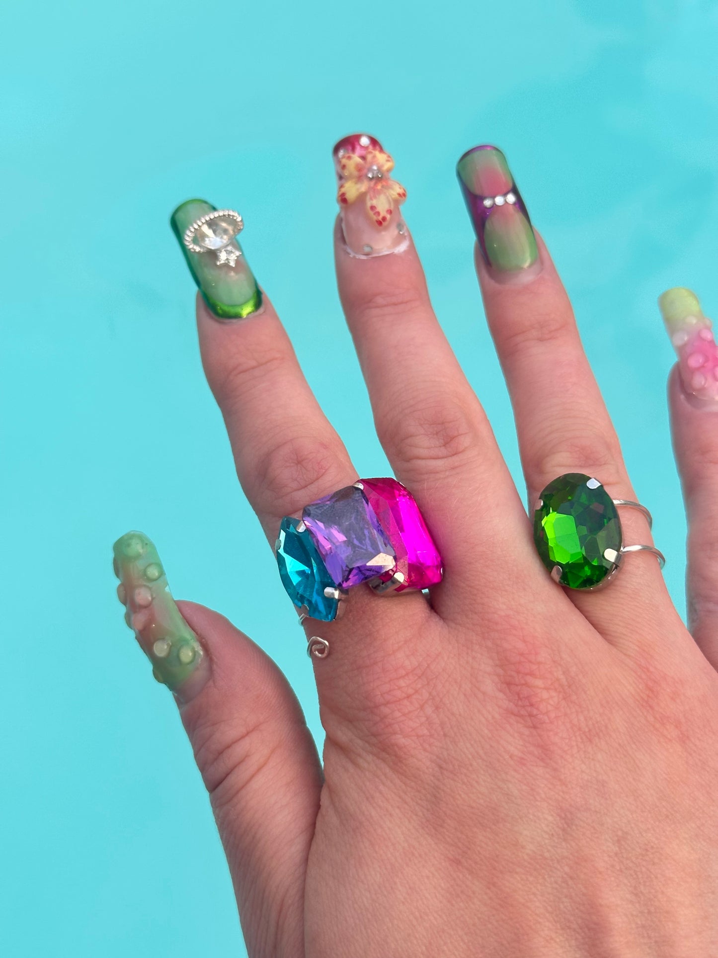 Hand with colorful gemstone rings and gradient nail polish against a blue background