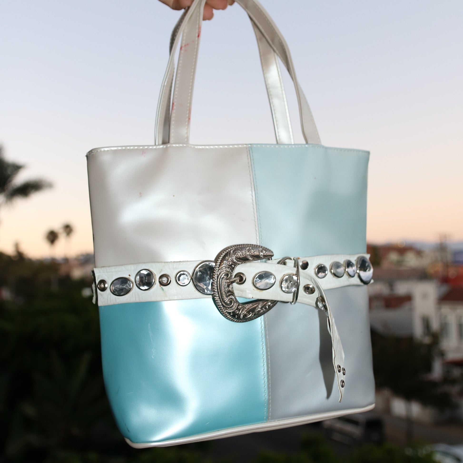 Hand holding a silver and blue handbag with decorative elements against a blurred background