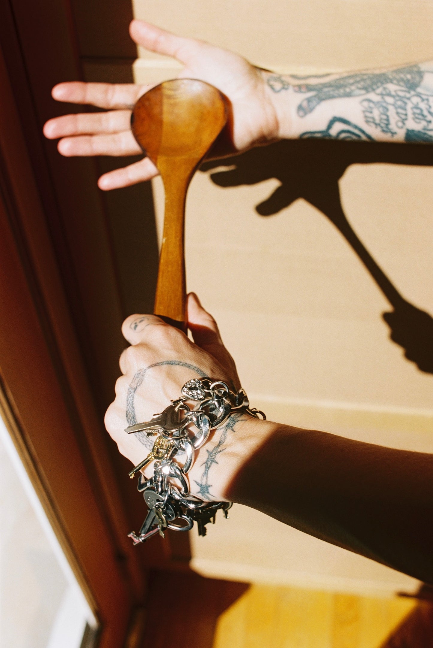 Hand holding a wooden spoon with tattoos and a bracelet, casting a shadow on a light-colored wall.